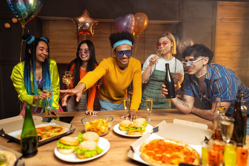 Group of friends making party at home on new year evening. Young women and men celebrating in the apartment drinking and eating, dancing and having fun. Concept about holidays and parties.