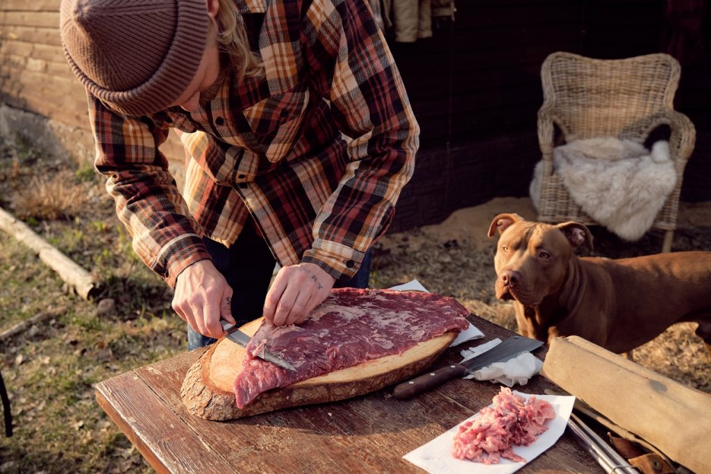 Young farmer cutting fresh meat for barbecue with his dog standing near by and watching for the process