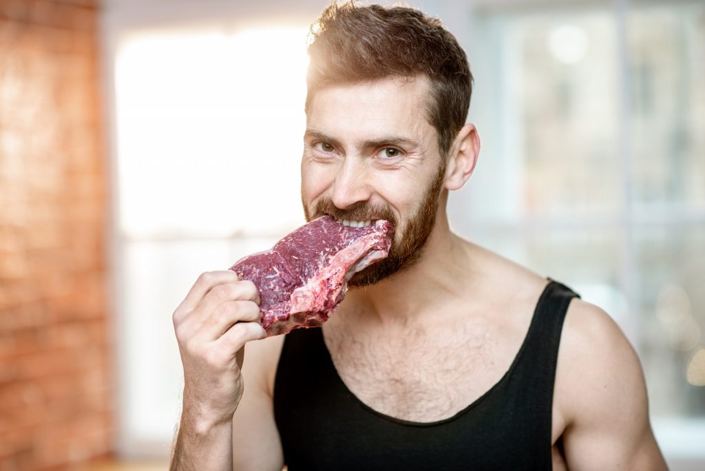 Portrait of a handsome sports man in black t-shirt biting raw meat steak indoors